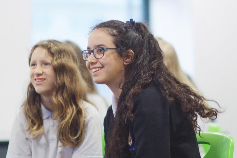 Two smiling young girls listen to an unseen speaker.