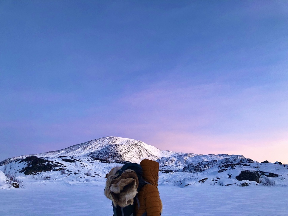 A person stood centre-frame with a fur blanket rolled up into their backpack. They are surrounded by snow covered ground and mountains. The sky is clear and blue, with pink hues resting at the top of the mountain