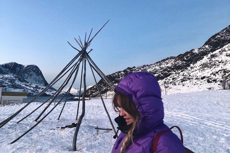 A person in a purple coat with the hood up, stood in front of a wooden tipi structure which is stood on a snowy landscape with dark mountains behind it