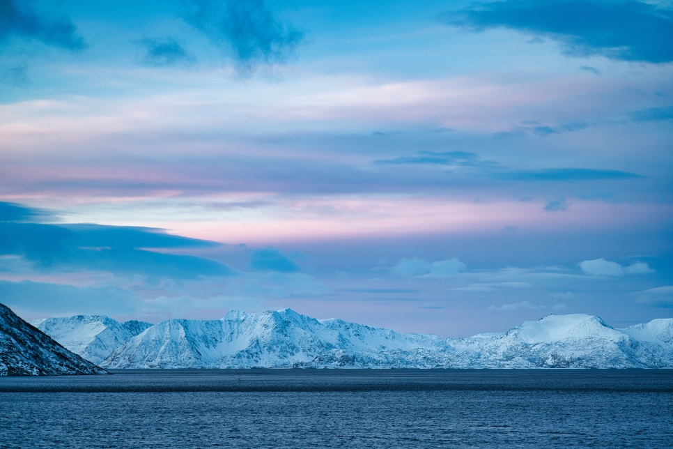 A dark lake in the foreground, with snow covered mountains across the background. The sky above is blue with some pink hues and there are clouds surrounding the outer edges of the image