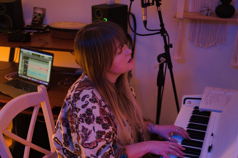 A person sat in a white chair playing a white keyboard/piano - they appear to be singing. Behind the is an open laptop and music recording software on the screen. They are surrounded by other recording equipment in the room.