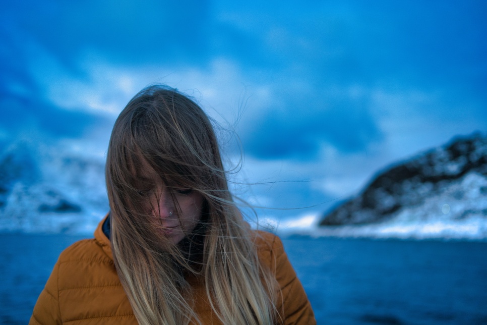A person stood in front of a vast dark lake with snow covered mountains either side. They're looking down and their hair is blowing in the wind, obscuring most of their face