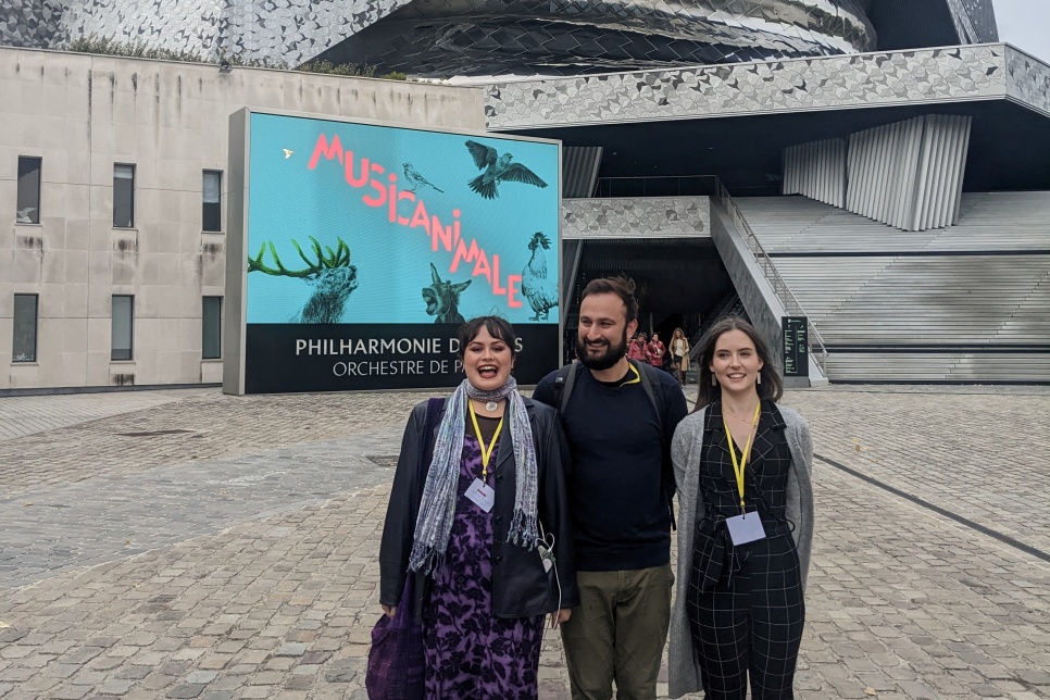Three people are stood together smiling in front of the Philharmonie De Paris building