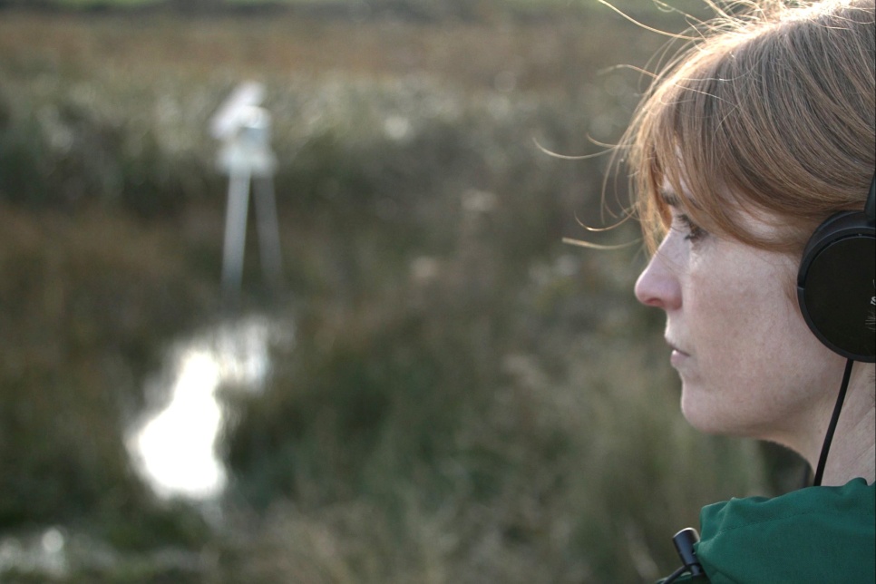 Female on right side of screen with headphones looking out over peatlands