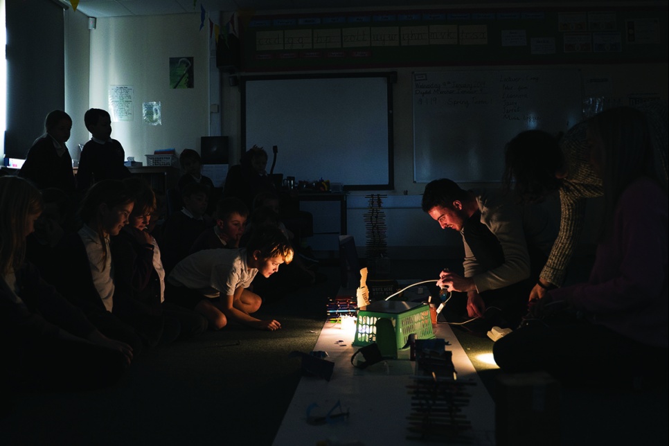 Teacher shows demonstrates a creative project on the floor to a group of children.