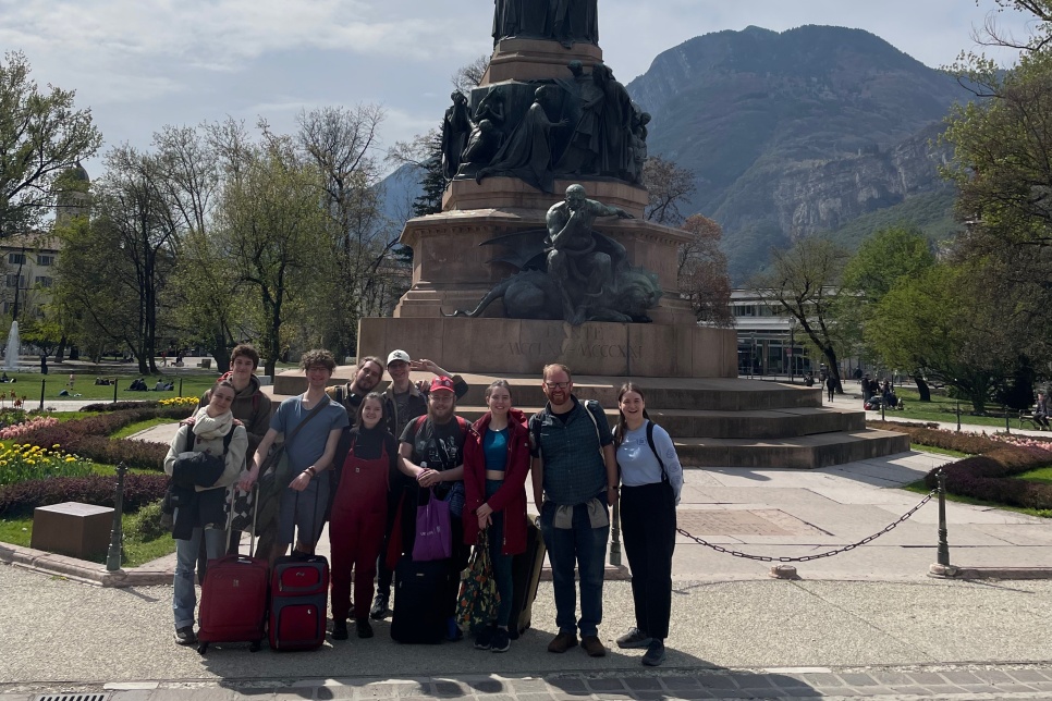 A group of young people are gathered in front of a statue. There are a few suitcases with them, and they are all smiling at the camera. Behind the large statue are trees and mountains.