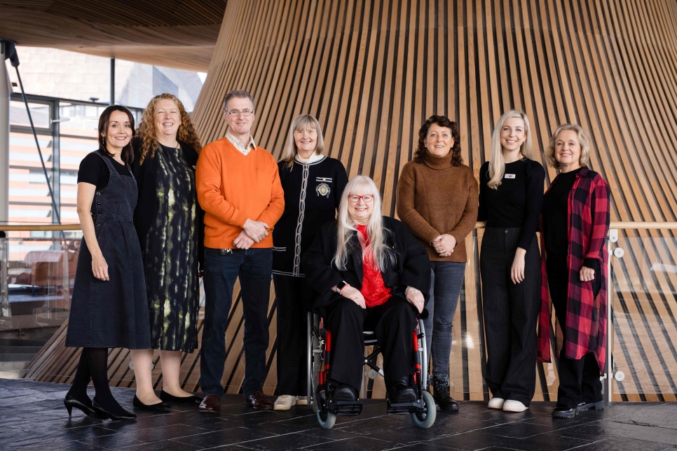 Group of seven adults, one in a wheelchair, posing together inside the Senedd buliding front of a wooden slatted wall.