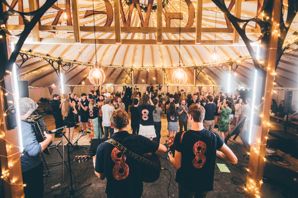  Musicians play to a dancing crowd in a festively lit tent, with string lights and a warm, communal atmosphere.