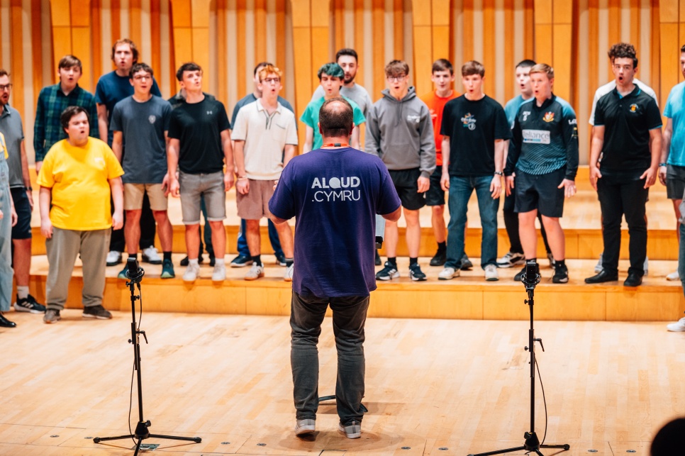 ALOUD Academi rehearsal 2025 A man with a purple t-shirt that says ALOUD CYMRU on the back, is stood in front of a large group of a choir of boys in a wooden hall