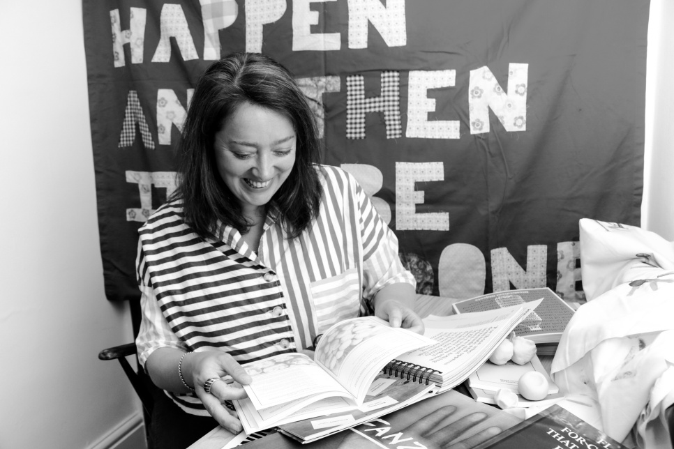 Clare Charles_photo by Phoebe Davies A woman in a striped shirt smiling at a book, in black and white