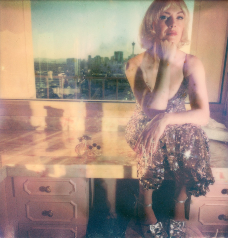 GWENNO-UTOPIA Women with short blonde bobbed hair, sat in a sparkling dress in front of a sunny cityscape.
