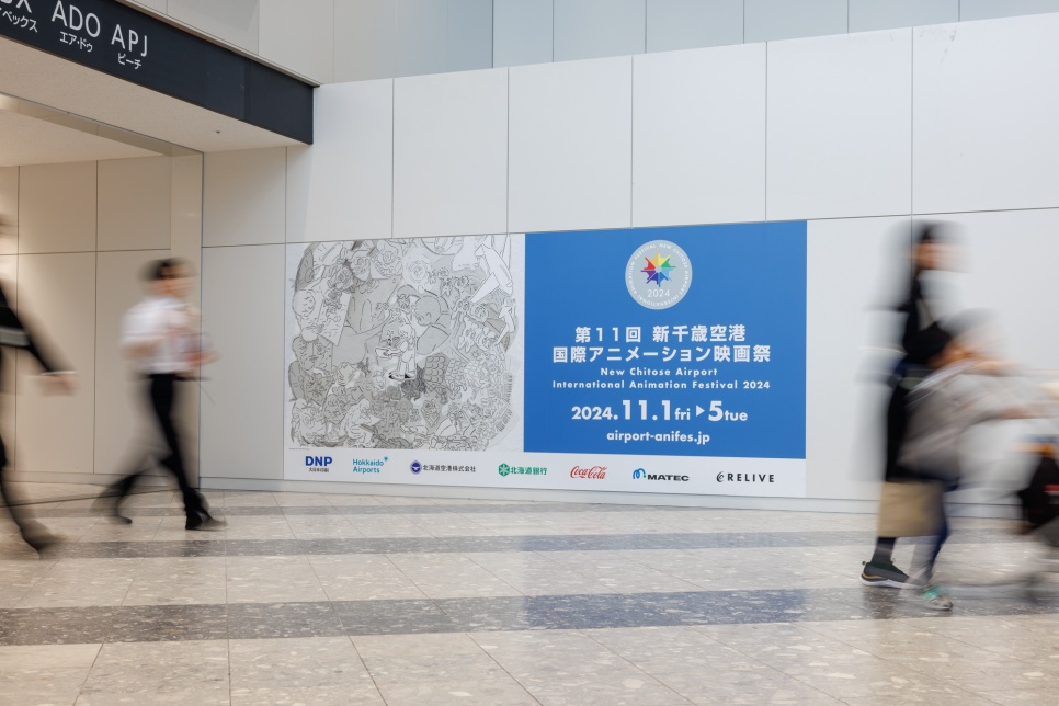 New Chitose Airport International Animation Festival People walking in a white corridor by a large blue, white and black advertisement for an animation festival in Japanese text.