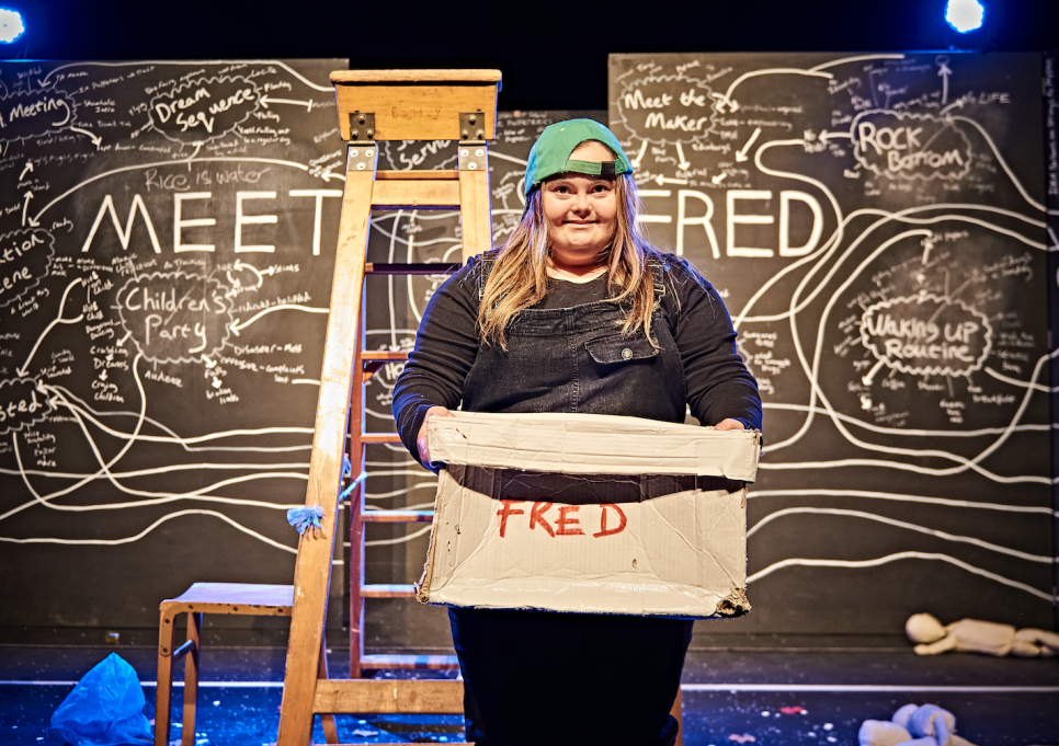 HIJINX - MEET FRED - credit - Jorge Lizalde A woman in a green baseball cap is stood smiling, holding a box, in front of a blackboard with chalk diagrams and the writing 'MEET FRED'
