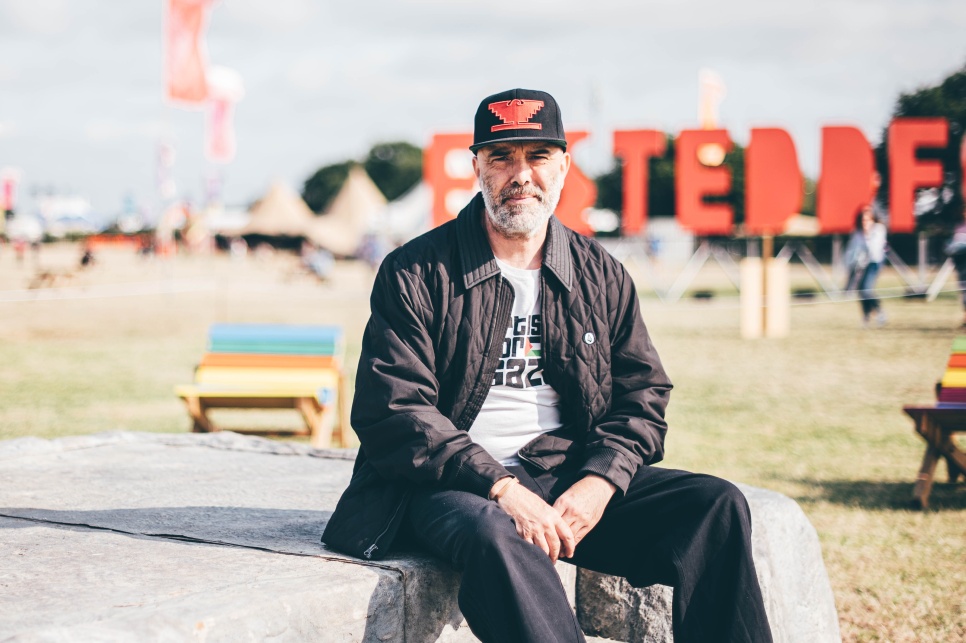 Cian Ciarán at the EISTEDDFOD 2025 Wrecsam - credit - Aled Llywelyn A man in a black jacket and black baseball cap sat on a large stone in front of a large red EISTEDDFOD sign