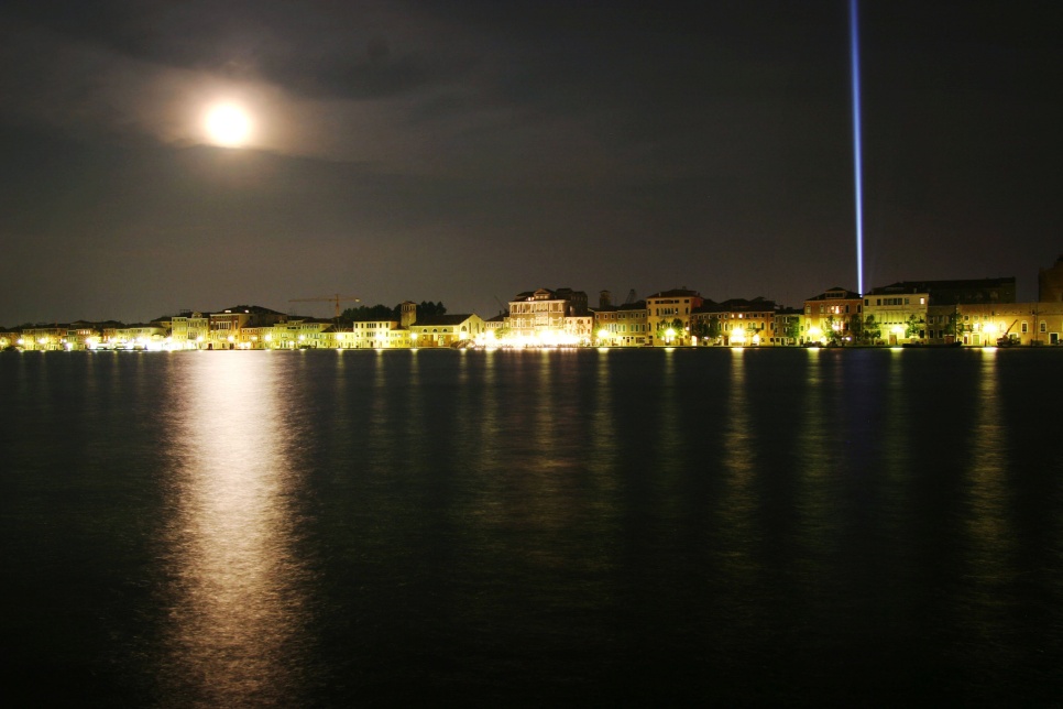 A night-time waterfront scene with buildings lit by warm yellow lights reflected on the water. A bright full moon glows in the cloudy sky, casting a silvery reflection on the water, while a tall vertical beam of blue-white light rises from the right side of the town into the night sky.